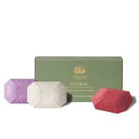 Three luxurious pampering Caswell - Massey floral-scented soaps in purple, white, and red in front of their green cardboard packaging box. The soaps and the box feature embossed detailing.