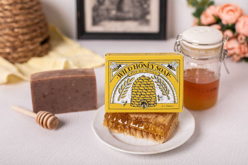 A natural skincare setup featuring a bar of honey soap labeled "Primitive House Farm's Wild Honey Soap" next to a honeycomb piece, a dipper on a jar of honey, with pink roses and a