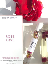 An image of a Hydra Bloom Beauty Rose Love Essential Perfume Roll-on Oil in a clear bottle with rose petals inside, next to its packaging and a vibrant red rose under bright lighting.