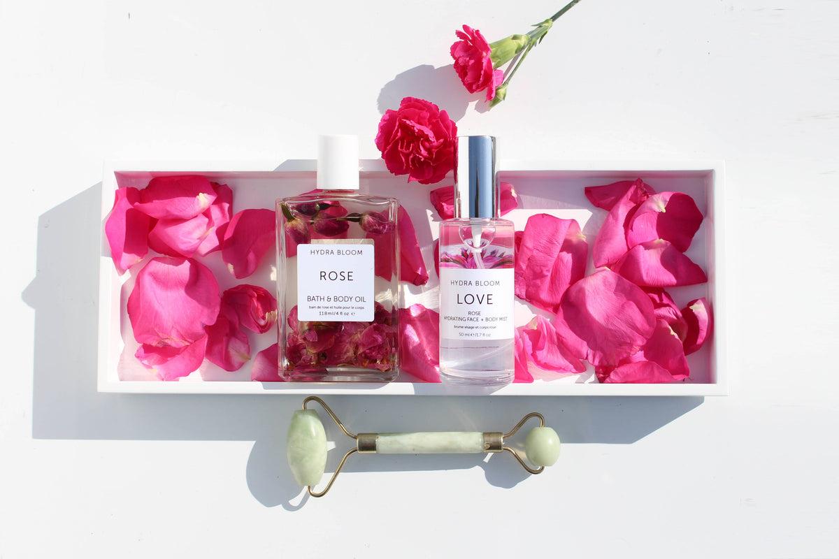 A tray with Hydra Bloom Beauty Rose Love Bath and Body Oil, surrounded by pink rose petals. Includes a jade roller, perfume, and lotion bottles under bright sunlight.