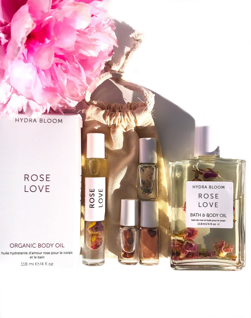 A collection of Hydra Bloom Beauty Rose Love Bath and Body Oil, including organic body oil, bath oil, and small roller bottles enriched with rose essential oils, displayed alongside pink flowers on a bright background.