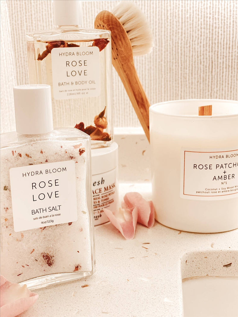 A cozy bathroom setting featuring Hydra Bloom Beauty Rose Love bath products including the face and body mist, body oil, and a candle infused with Australian Flower Essences, alongside a brush and petals, all bathed in warm.