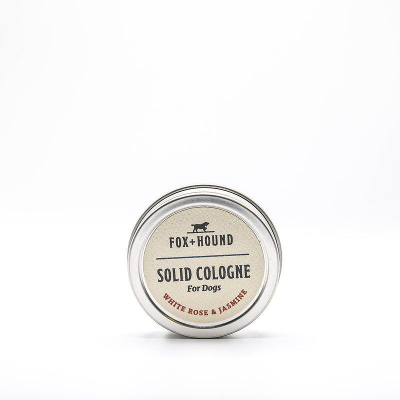 A small circular tin labeled "Fox + Hound White Rose and Jasmine Solid Cologne" is centered on a plain white background. The solid cologne's label features the scent description "White Rose + Jasmine." The tin has a metallic finish and boasts a simple, clean design.