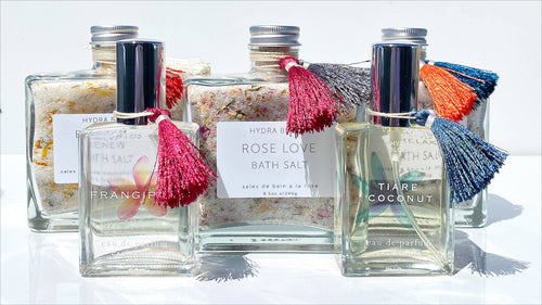 Five glass bottles of Hydra Bloom Beauty Rose Love Bath Salts and perfume labeled "frangipani," "rose love," and "tiare coconut," with colorful tassels, displayed against a bright white background. One of