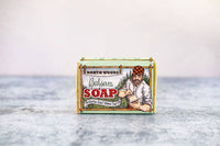 A bar of Primitive House Farm Balsam Soap in a colorful cardboard box featuring an illustration of a bearded man in a plaid shirt on a gray background. This cold-process soap captures the essence of