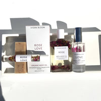 A collection of "Hydra Bloom Beauty" branded skin care products, including Rose Love organic body oil in a bottle and roller, a bar of soap, and bath oil with floating petals, arranged under natural.