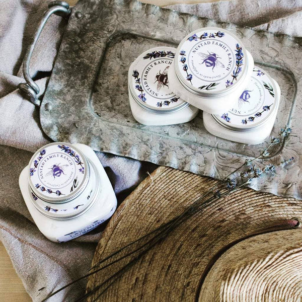 A collection of porcelain jars with vintage blue floral design, labeled "Nustad Family Ranch French Lavender Shea Body Butter," arranged on a rustic grey tray infused with lavender essential oil, featuring wooden and floral accents.