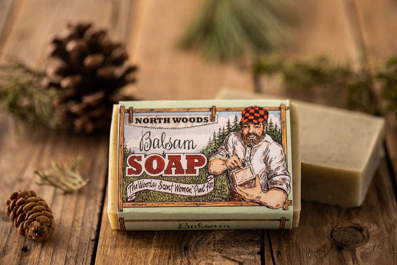 A bar of Primitive House Farm Balsam Soap in a decorative box featuring a lumberjack illustration, placed on a wooden table with pine cones and pine branches nearby.