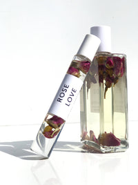 A clear bottle of Hydra Bloom Beauty Rose Love Essential Perfume Roll-on Oil with pink rose petals inside, alongside a matching rollerball application tube, both set against a plain white background.