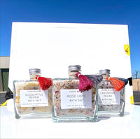 Three glass jars of organic bath salts labeled "eucalyptus renew," "Hydra Bloom Beauty Rose Love Bath Salts," and "lavender relax" from Hydra Bloom Beauty, adorned with colorful tassels, displayed against a