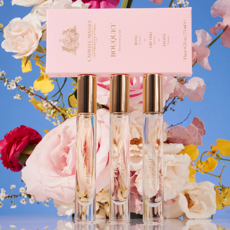 Three elegant Caswell - Massey Bouquet Discovery Set perfume bottles with a pink label in front, surrounded by a vibrant arrangement of roses and yellow flowers from the New York Botanical Garden, against a blue background.
