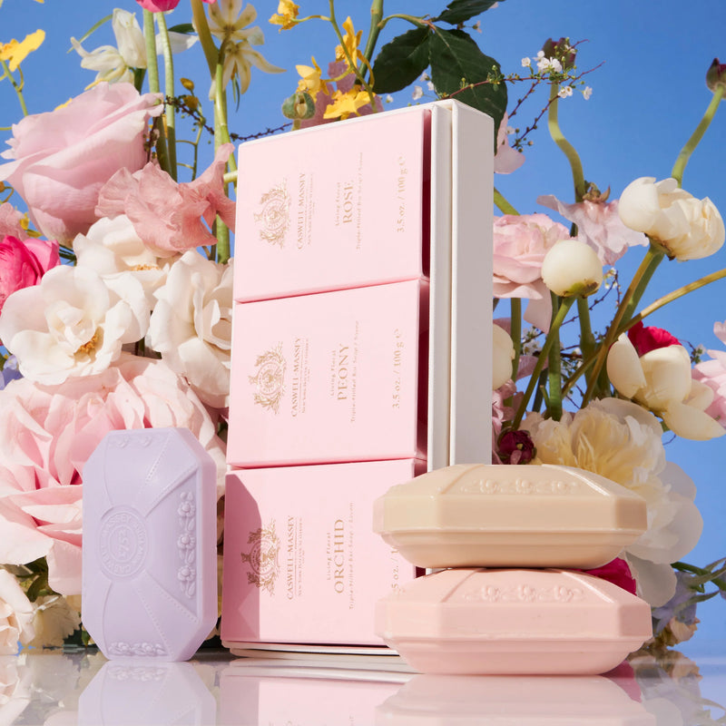 Three pink, luxurious Caswell Massey soap bars and their floral-patterned boxes arranged amidst a backdrop of lush flowers under a blue sky.
