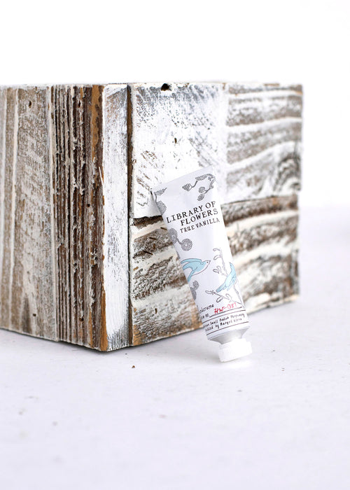 A hand cream tube labeled "Library of Flowers True Vanilla Petite Treat Handcreme" by Margot Elena leans against a rustic, weathered wooden block with peeling white paint, set against a plain white background.