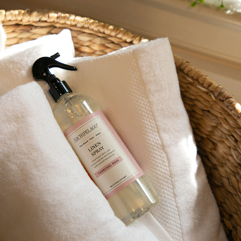A bottle of Archipelago Charcoal Rose Linen Spray resting on a white towel inside a wicker basket, in a cozy, sunlit setting.