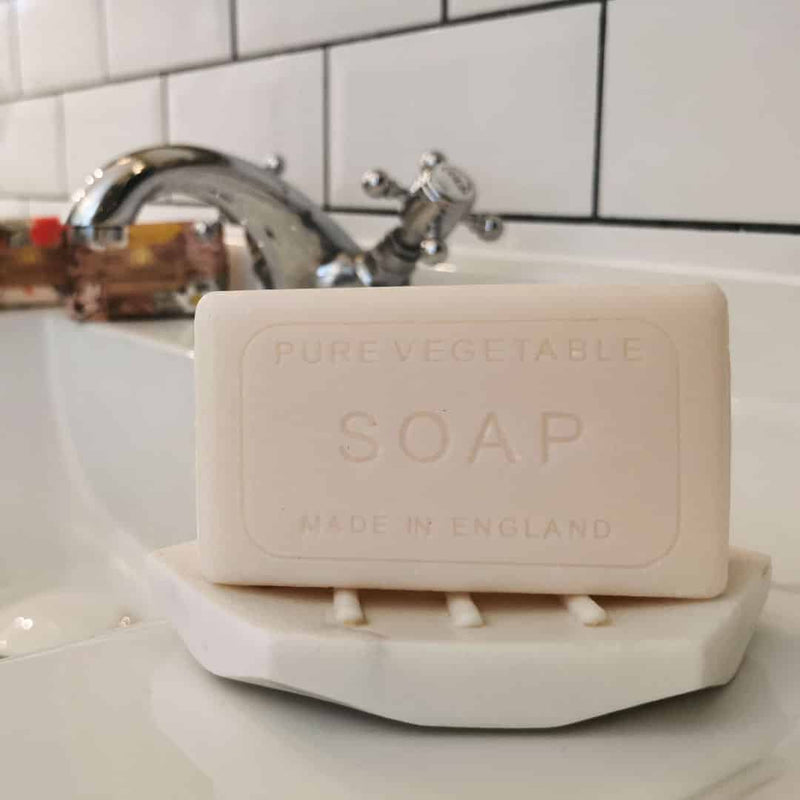 A bar of "The English Soap Co. Anniversary Indian Sandalwood Soap" made in England, resting on a white soap dish beside a sink with a shiny faucet, against a white tiled background.