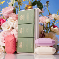 A stack of elegantly packaged, triple-milled Caswell Massey Three-Soap Sets in various pastel shades, displayed against a backdrop of lush pink and white roses under a clear blue sky.
