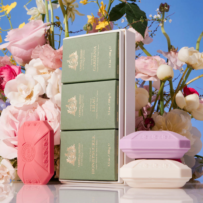 A stack of elegantly packaged, triple-milled Caswell Massey Three-Soap Sets in various pastel shades, displayed against a backdrop of lush pink and white roses under a clear blue sky.