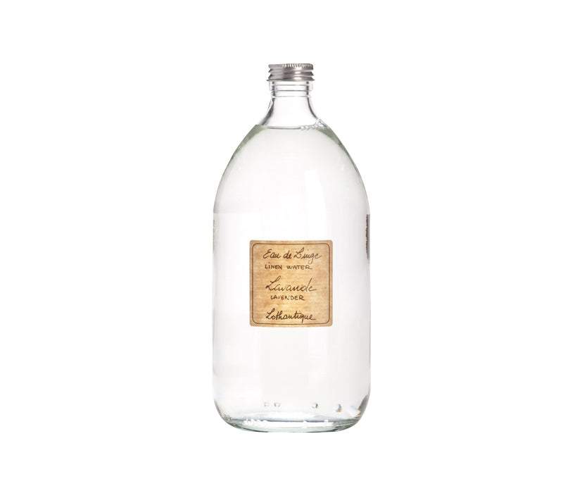 A clear glass bottle with a silver cap, filled with a transparent liquid. It has a beige label affixed to the front, featuring text in a stylish font that reads "Lothantique Lavender Linen Water Spray - 1 liter/33oz." This scented water is perfect for use with your steam iron to refresh linens. The bottle is placed against a white background.