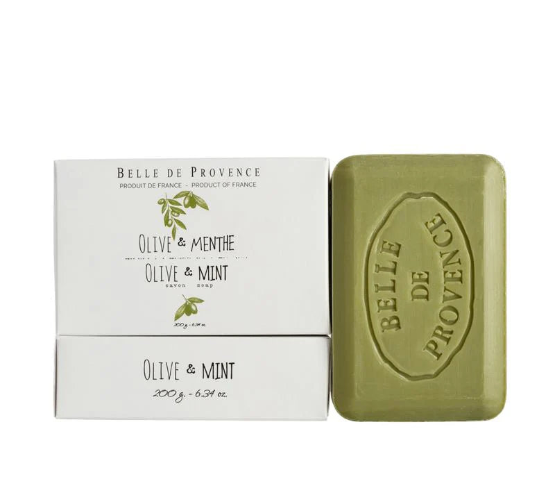 A Lothantique Belle de Provence Olive & Mint 200gm Soap is placed next to two matching white boxes that are also labeled "Olive & Mint." The boxes feature minimalist artwork of aromatic herbs, showcasing olive and mint plants. The soap bar is olive green with an embossed brand name.