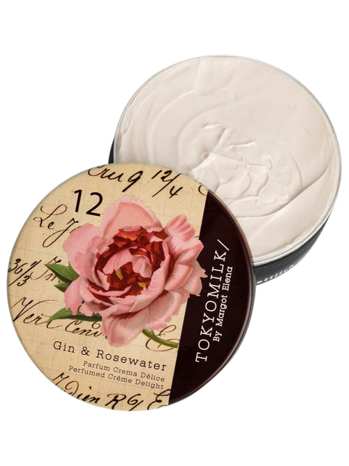 An open jar of Margot Elena's TokyoMilk Gin & Rosewater Parfum Crema Délice with a floral and vintage-designed lid lying next to it, featuring a pink rose and script elements on a brown.