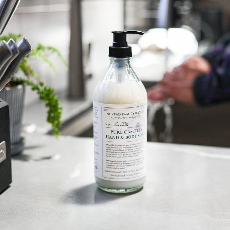 A bottle of Nustad Family Ranch Wild Magnolia Pure Castile Hand & Body Soap with a pump dispenser, placed on a kitchen counter near a sink with a blurred person in the background.