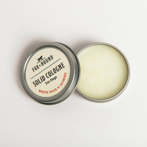 A small round tin of travel-friendly Fox + Hound White Rose and Jasmine Solid Cologne, labeled as White Rose + Jasmine scent. The tin is open, revealing the smooth, off-white dog solid cologne inside. The tin lid is placed next to the container on a white background.