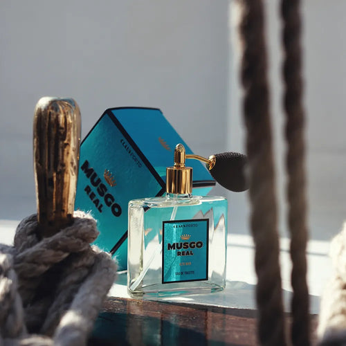 A bottle of Claus Porto Musgo Real Alto Mar Eau de Toilette with a luxurious aroma is placed in front of its blue packaging, with part of the image framed by thick coiled ropes on a reflective glass surface.