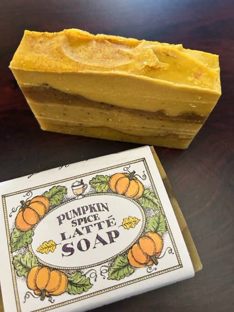 A bar of Primitive House Farm Pumpkin Spice Latte soap with a textured top rests above its labeled box on a wooden surface. The soap is yellow-orange with visible layers and infused with coffee extract.