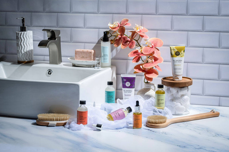 A modern bathroom sink surrounded by various beauty and skincare products, including Woolzies Nourishing Vanilla Hand Cream, a soap bar, wooden brushes, and a vibrant orchid on a marble countertop.
