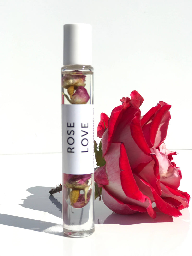 A roller bottle of rose-scented oil labeled "Hydra Bloom Beauty Rose Love Essential Perfume Roll-on Oil," adorned with dried rose buds inside, standing next to a vibrant pink and white rose against a white background, is infused with organic floral.