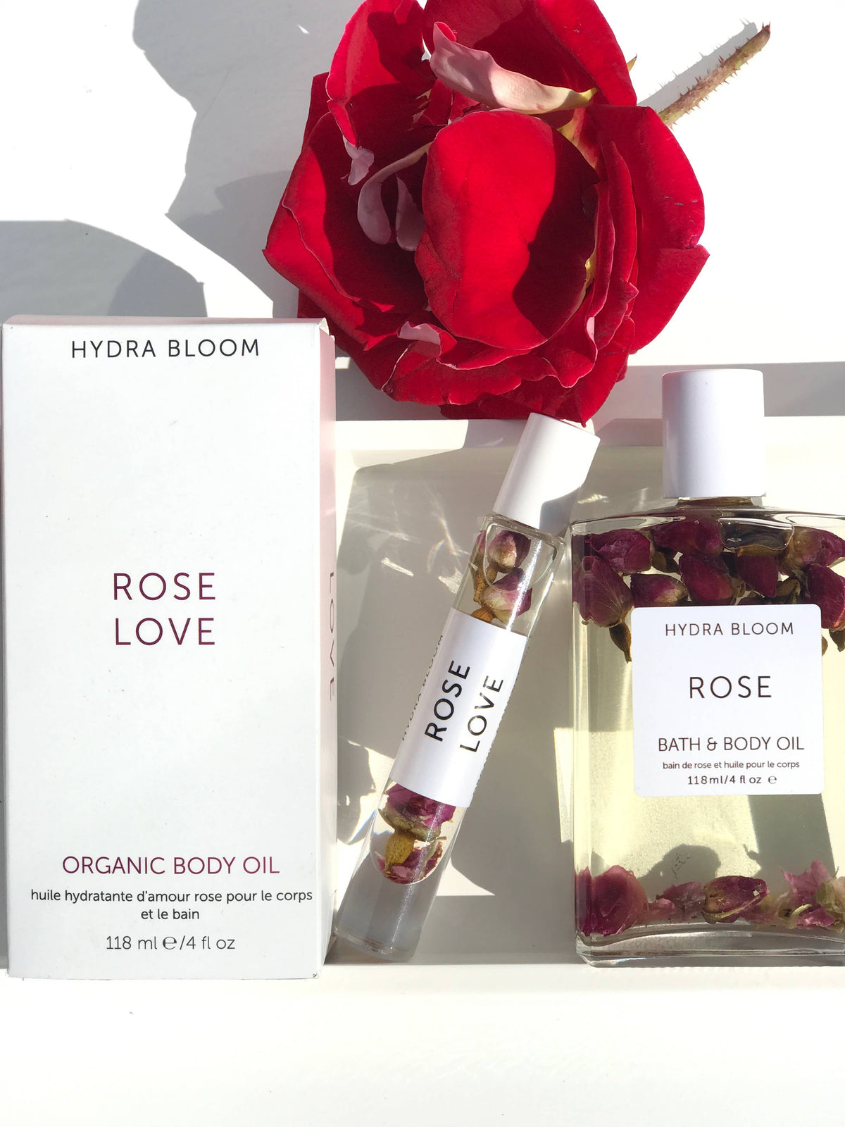 An arrangement featuring a red rose, a bottle and a smaller roll-on bottle of Hydra Bloom Beauty Rose Love Bath and Body Oil, and a white box labeled accordingly, all placed on a white
