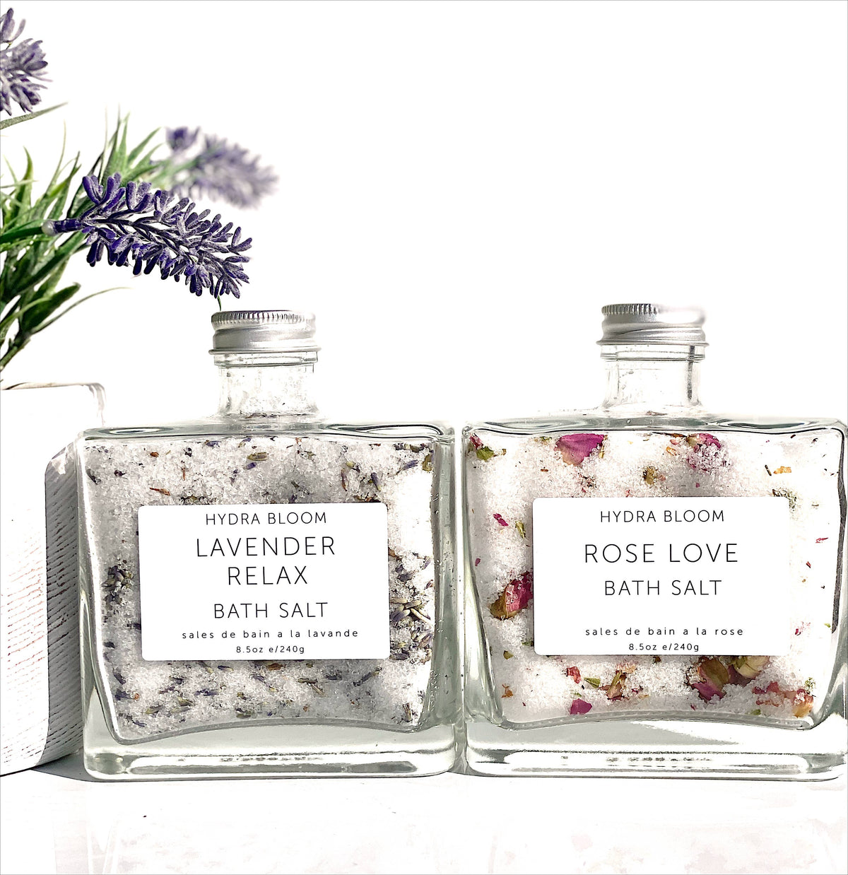 Two square glass bottles of Hydra Bloom Beauty Rose Love Bath Salts labeled "lavender relax" and "rose Epsom salt," filled with scented salts and visible botanical pieces, set against a white background.