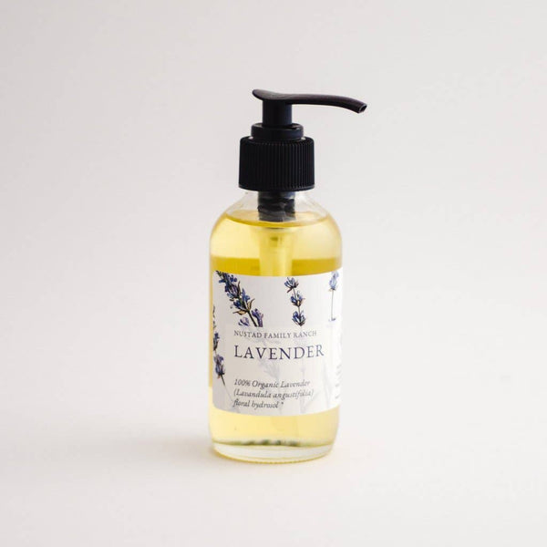 A clear bottle of Nustad Family Ranch French Lavender Body Oil with a black pump, labeled "nustad family ranch lavender," specifying "100% organic French lavender essential oil soap" on a plain white background.