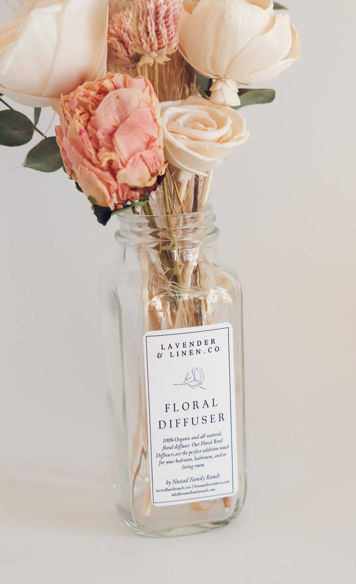 A Nustad Family Ranch Tuberose & Honey reed diffuser in a clear glass bottle with a label that reads "Nustad Family Ranch Tuberose & Honey floral diffuser" surrounded by pink peonies and pampas against a light background.