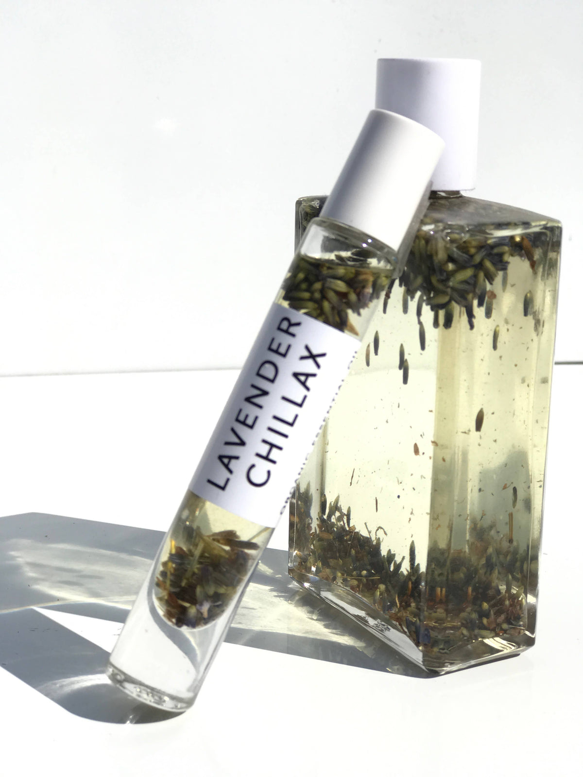 A clear glass bottle containing Hydra Bloom Beauty Lavender Chillax Organic Essential Perfume oil with lavender blossoms, labeled "lavender chillax," stands against a white background, with sunlight casting its shadow.