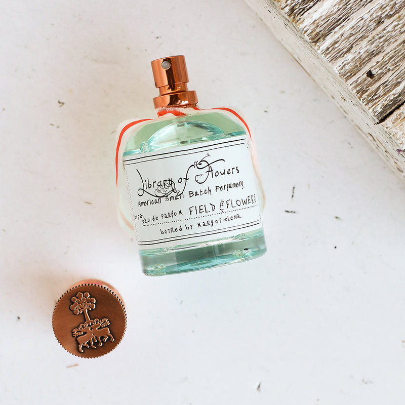 A square perfume bottle with a copper spray top and clear blue liquid labeled "Margot Elena, Library of Flowers Field & Flowers Eau de Parfum" rests on a white rustic surface next to a round copper cap with a floral.