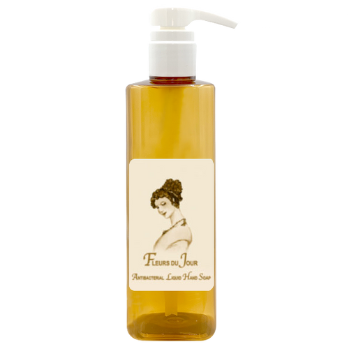 A transparent pump bottle containing amber-colored La Bouquetiere Fleurs du Jour/Marina Blue antibacterial liquid hand soap with a label featuring an illustration of a woman and the text "fleurs du jour - aromatherapy olive hair soap".