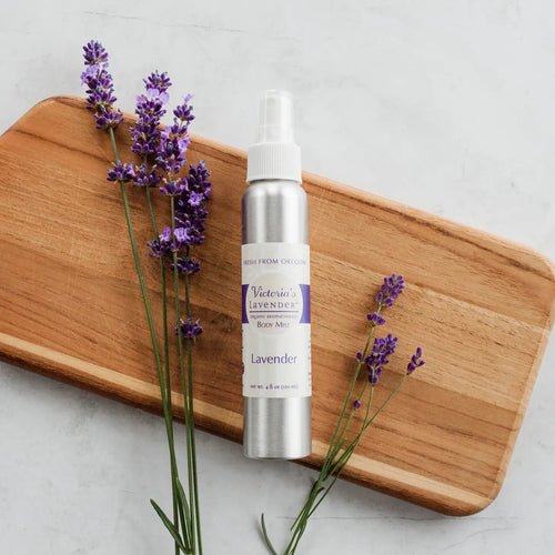 A bottle of Victoria's Lavender Aromatherapy Body Mist placed on a wooden board alongside fresh lavender sprigs and aloe vera against a marble background.
