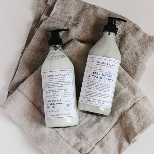 Two bottles of Nustad Family Ranch French Lavender Pure Castile Hand & Body Soap on a cloth napkin. One bottle is shea butter hand lotion and the other is eco-friendly French Lavender Pure Castile Liquid Soap.