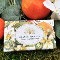 A bar of The English Soap Co. Orange Blossom Vintage Wrapped Soap, displayed on a green leafy background with orange and white flowers nearby.