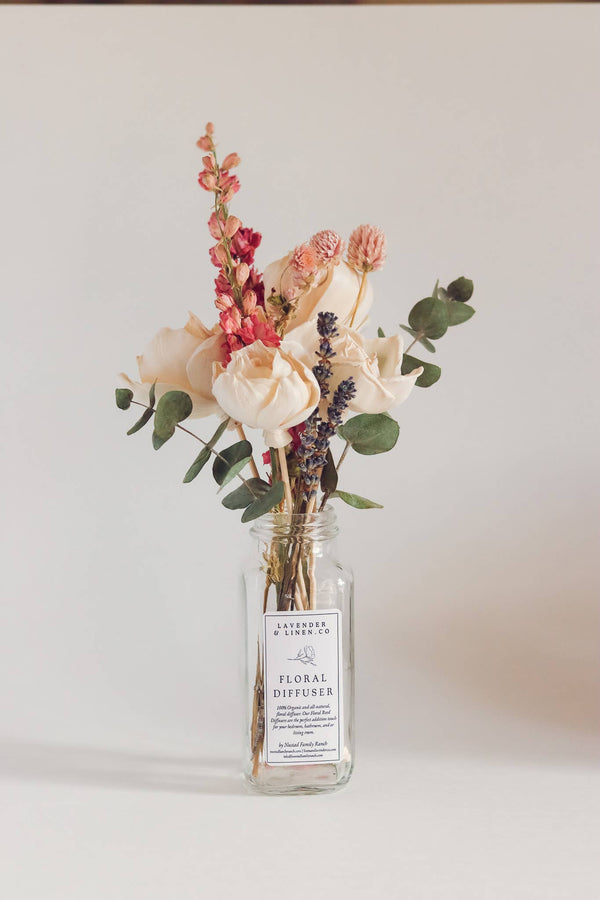 A Nustad Family Ranch French Lavender reed diffuser in a clear glass bottle labeled "floral diffuser," adorned with an arrangement of roses, eucalyptus, and various other flowers and plants, set against a