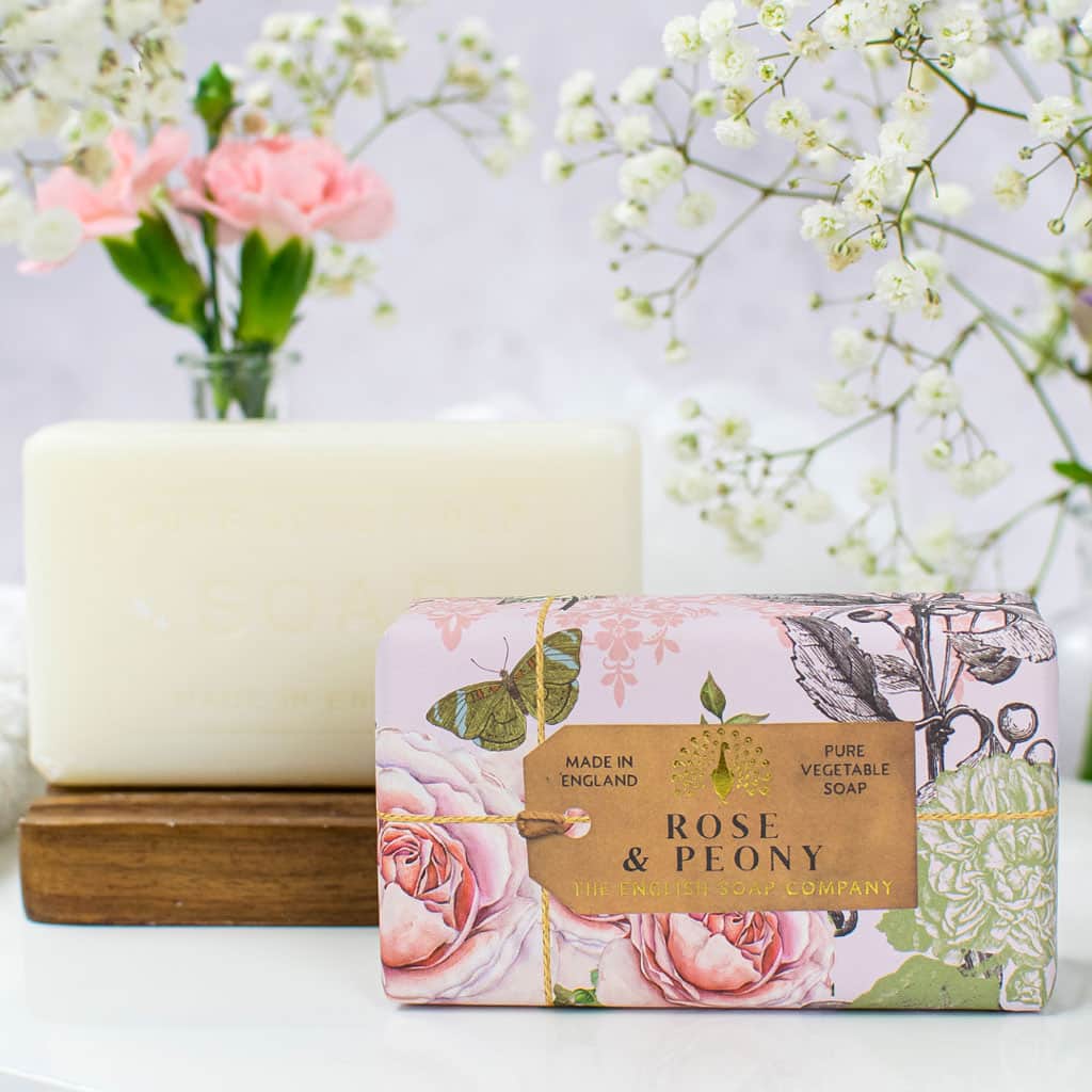 A bar of The English Soap Co. Anniversary Rose & Peony Soap rests on a wooden holder, accompanied by decorative packaging labeled "rose & peony," next to a vase of pink and white flowers, against a softly blurred background.