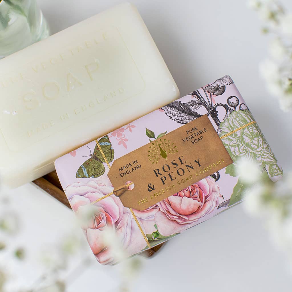 A bar of shea butter soap next to its floral packaging labeled "The English Soap Co. Anniversary Rose & Peony Soap" with white flowers around, on a light background. The soap packaging indicates it is made in The English Soap Co.