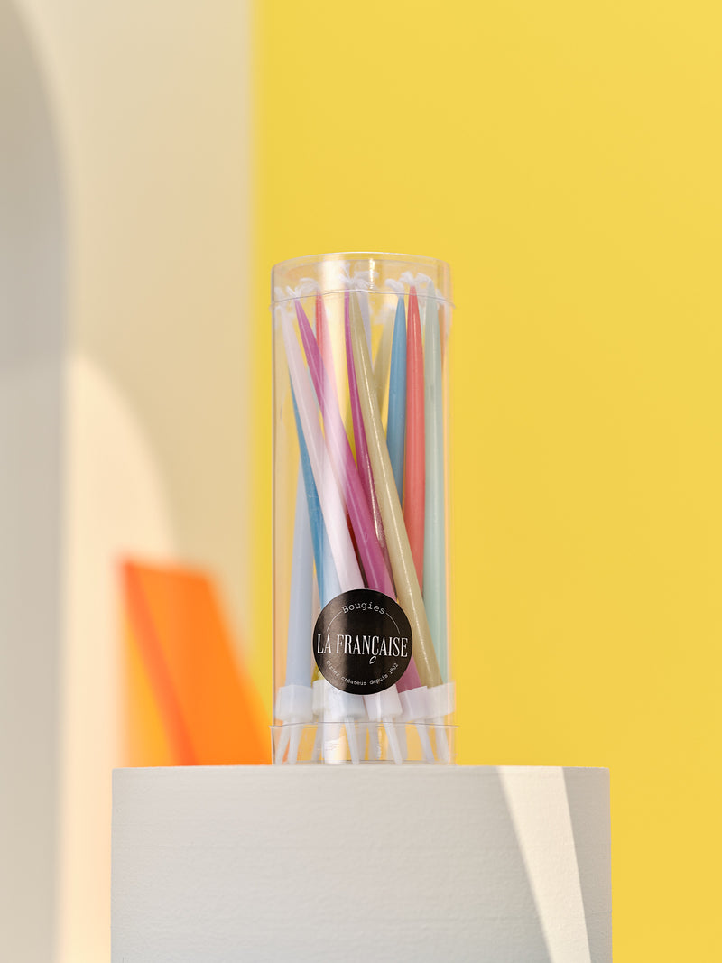 A clear glass container filled with Bougies La Francaise Twenty Multi-Colored Birthday Candles, displayed on a white pedestal against a yellow background with a partial view of orange elements.