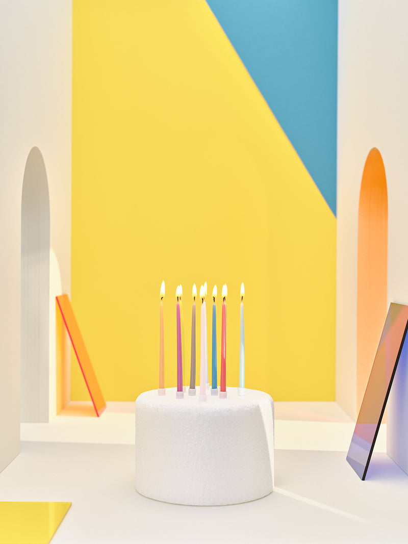 A white, round cake with Bougies La Francaise twenty multi-colored birthday candles in a geometric room with yellow and blue walls and abstract shapes.