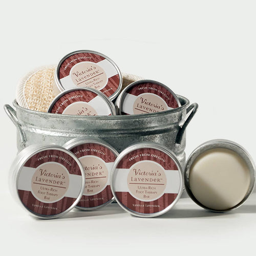 A collection of six labeled Victoria's Lavender Vanilla Lavender Foot Therapy Lotion Bars in a metal bucket, featuring creams, a lotion bar, and a body scrub, with a natural sponge on top.