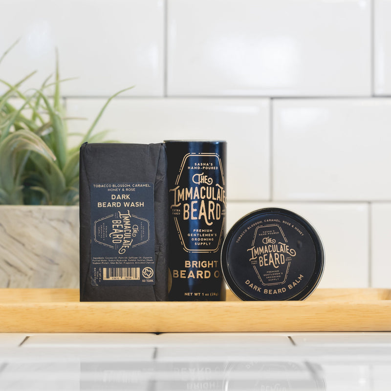 Men's grooming products, including a bar of soap, The Immaculate Beard - Beard Balm-Made in Texas - DUSK with a sandalwood vanilla scent, and beard wax, are displayed on a wooden shelf against a white tiled background.