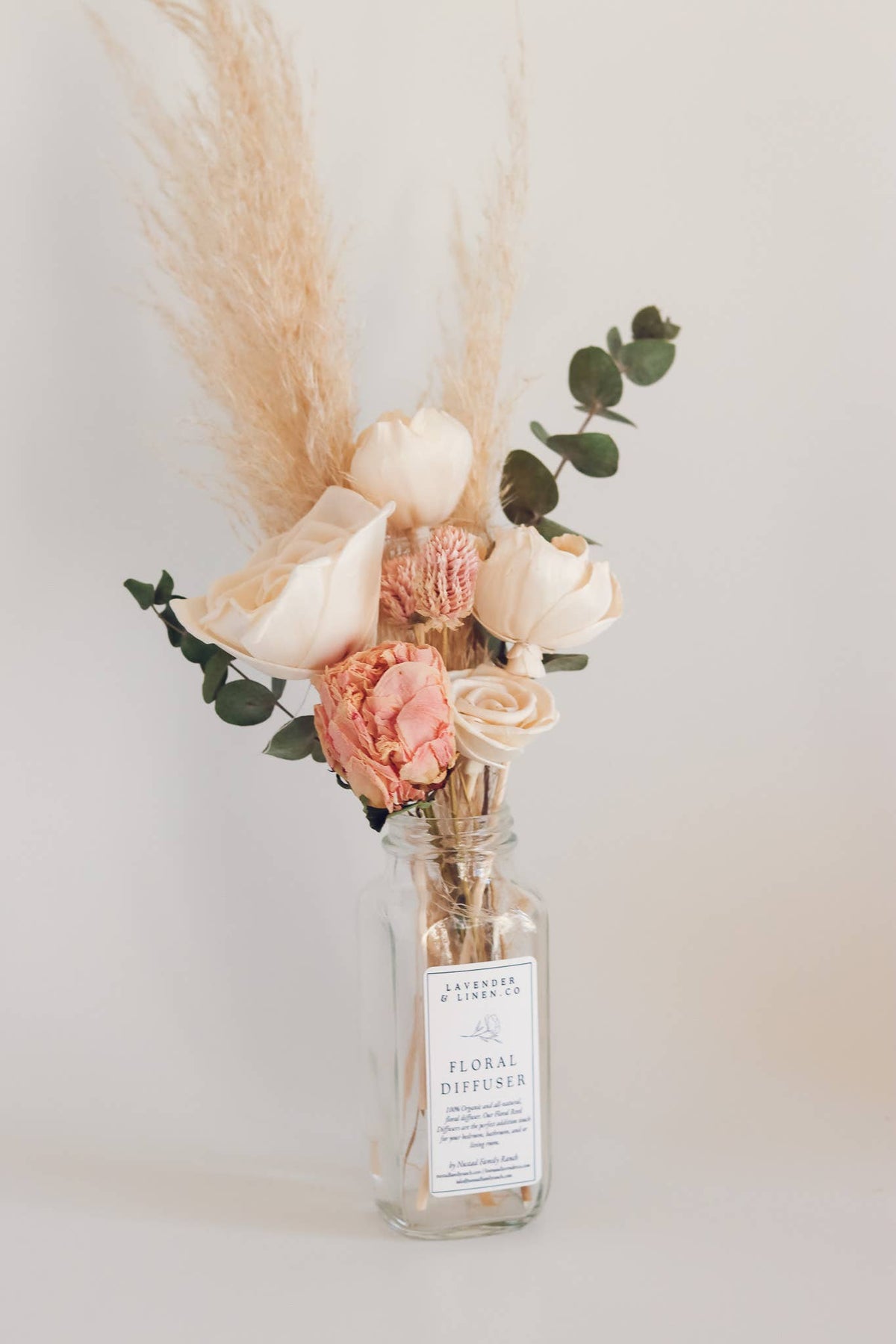 A Nustad Family Ranch Tuberose & Honey reed diffuser in a clear glass bottle with a label, surrounded by dried flowers such as pink peonies and pampas grass against a light beige background.