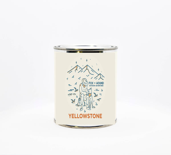 A Fox + Hound Yellowstone National Parks Wyoming soy candle in a small tin with a label depicting a wilderness scene featuring a fox, a hound, and a cabin under a mountain range, labeled "Yellowstone National Park.