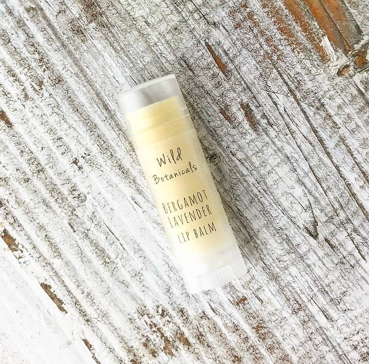 A tube of Wild Botanicals Bergamot Lavender Lip Balm lying on a rustic, weathered wooden surface. The lip balm packaging is simple and predominantly white.